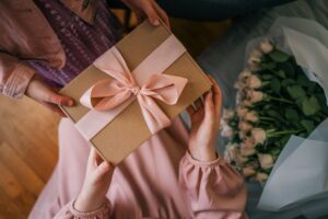 Frau überreicht ein persönliches Geburtstagsgeschenk mit rosa Schleife, daneben liegt ein Blumenstrauß mit hellen Rosen